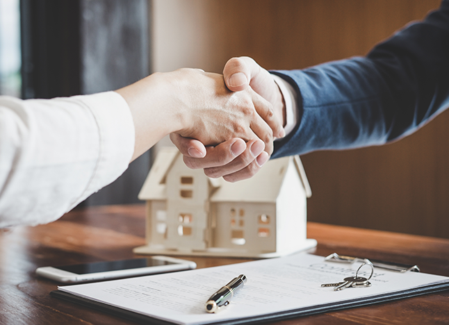 Themenbild Vermietung zu sehen sind zwei Hände, die geschüttelt werden, dahinter Vertragsunterlagen und ein Dekohaus auf einem Tisch.